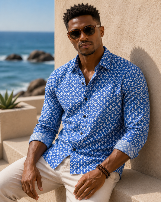 Man wearing a blue patterned shirt sitting by the ocean.
