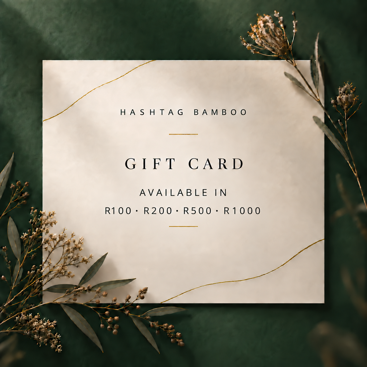 A square gift card with a warm cream background and elegant gold decorative lines, centered on a dark green textured backdrop with dried botanical sprigs. The card reads 'HASHTAG BAMBOO' and 'GIFT CARD' with denomination options R100, R200, R500, and R1000.