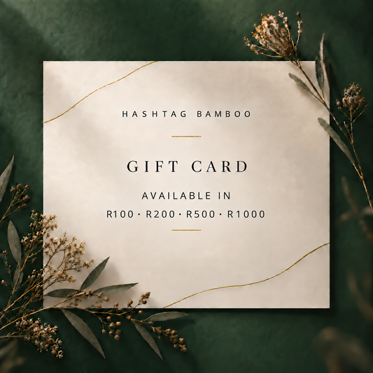 A square gift card with a warm cream background and elegant gold decorative lines, centered on a dark green textured backdrop with dried botanical sprigs. The card reads 'HASHTAG BAMBOO' and 'GIFT CARD' with denomination options R100, R200, R500, and R1000.