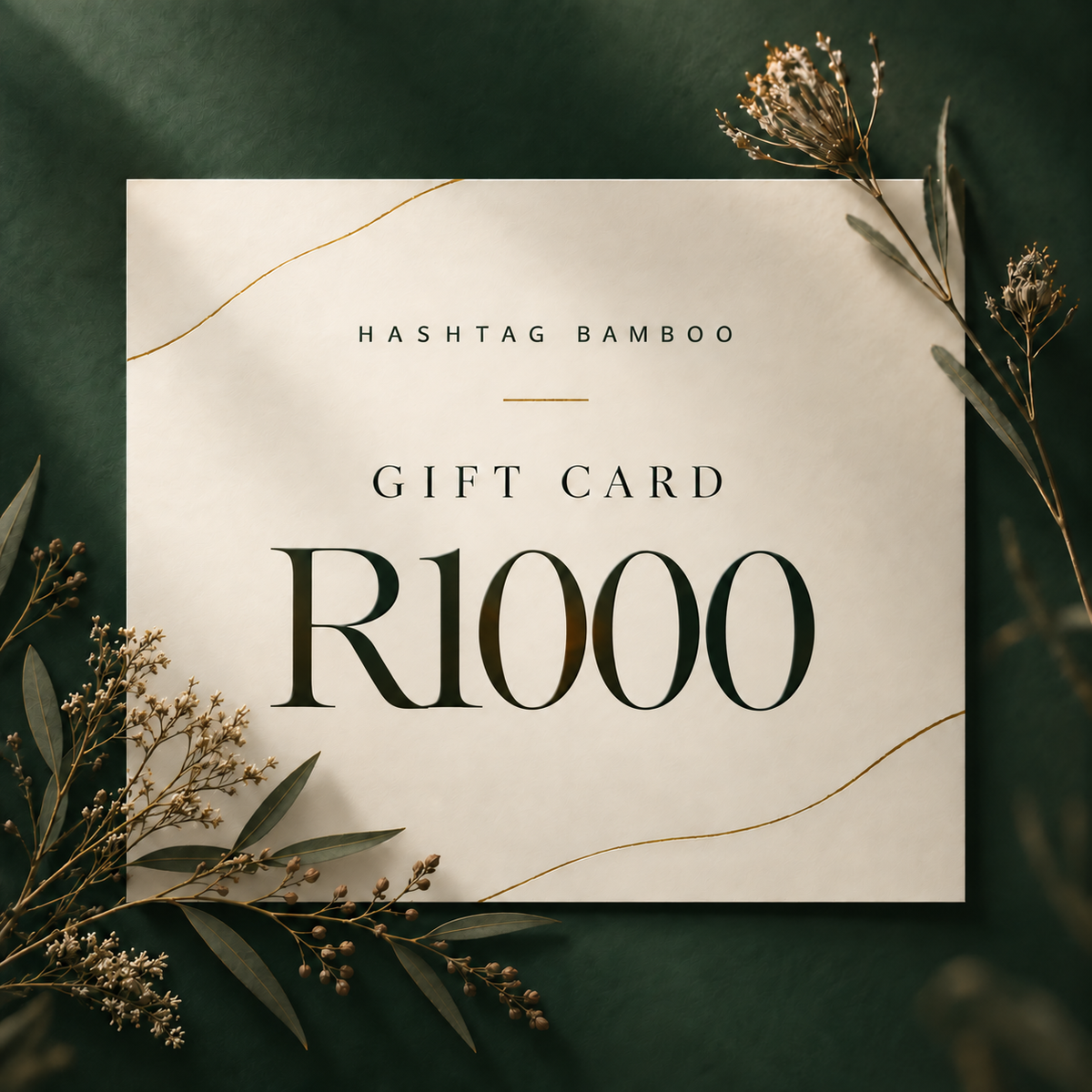 A square gift card with the text 'HASHTAG BAMBOO GIFT CARD R1000' in dark green lettering on a warm cream background, styled with dried botanicals on a dark green surface.