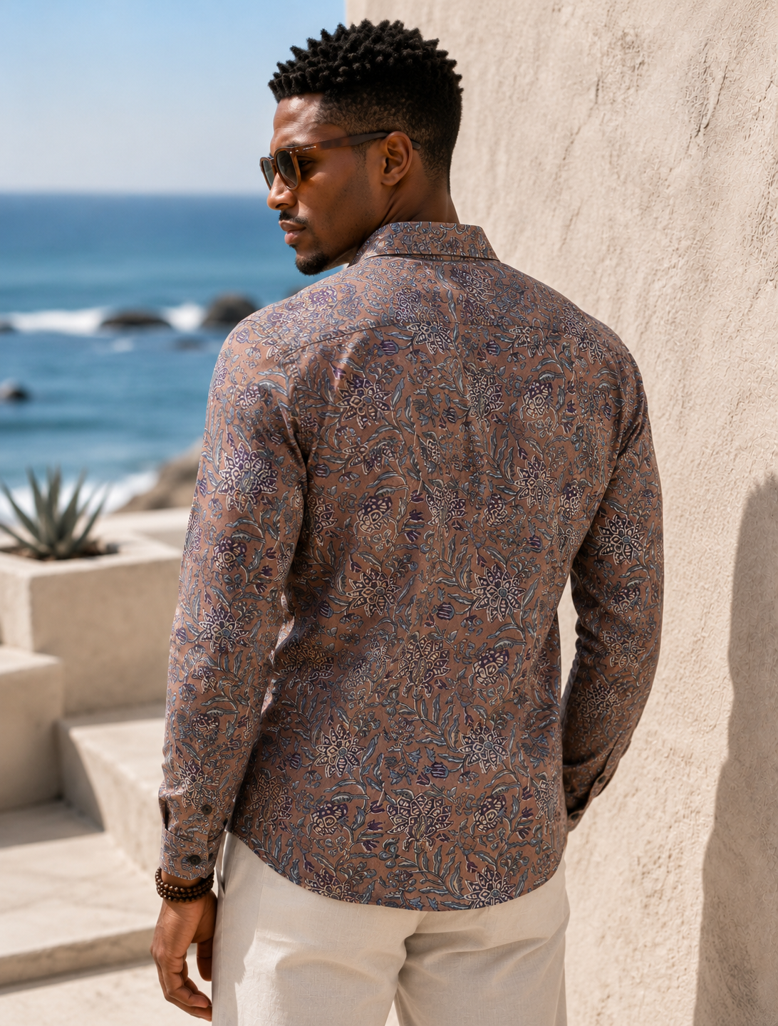 Man wearing a patterned shirt leaning against a stone wall with ocean view