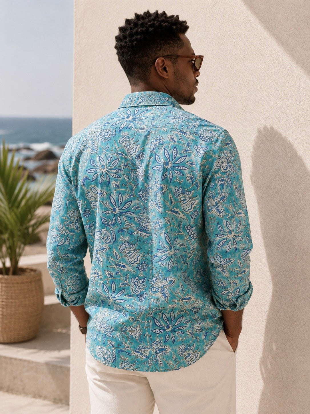 Man wearing a blue patterned shirt standing against a white wall with ocean view.